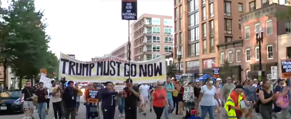 Crowd of demonstrators marching down a city street holding a large banner that reads “TRUMP MUST GO NOW.”