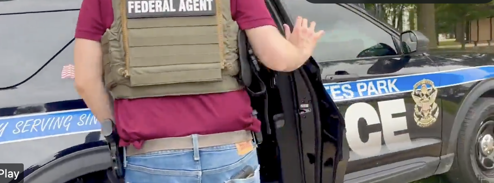Close-up of a federal agent wearing a tactical vest and standing beside a police vehicle with one hand raised.
