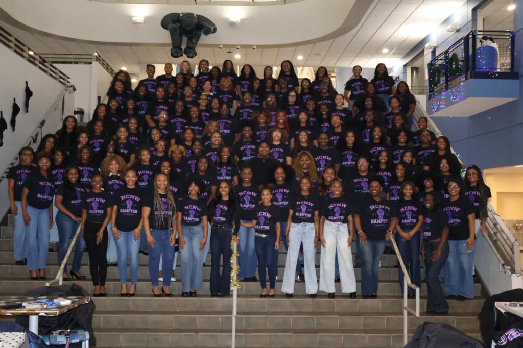 Large group photo of Hampton University’s NCNW chapter standing on staircase steps, all wearing matching black NCNW shirts with purple lettering and jeans, smiling toward the camera.