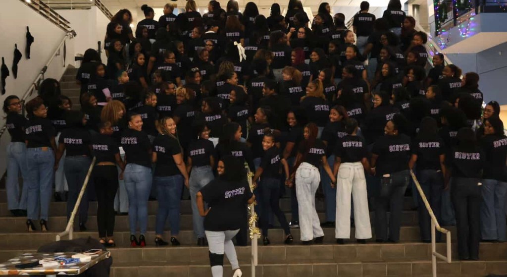 Back view of the Hampton University NCNW chapter on staircase steps, showing the word “SISTER” printed on the back of their matching black shirts as members turn, laugh, and pose together.