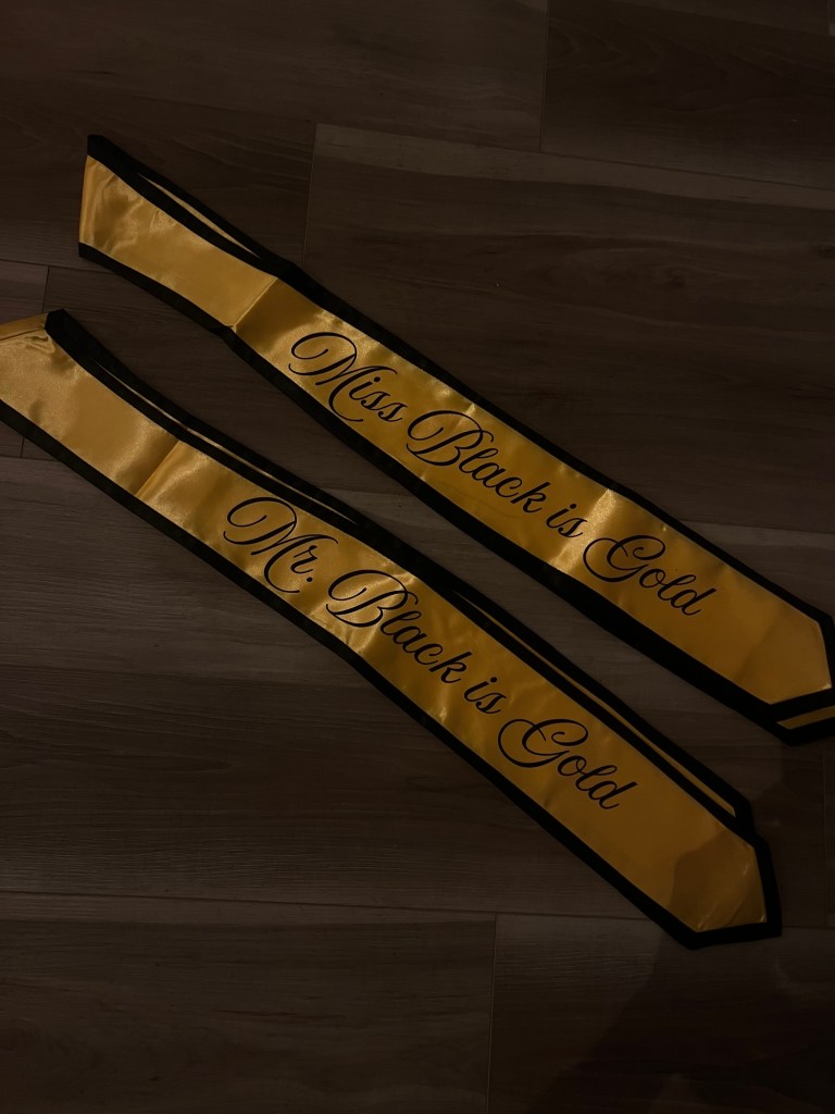 Two gold sashes with black trim with black cursive text "Miss/Mr. Black is Gold".