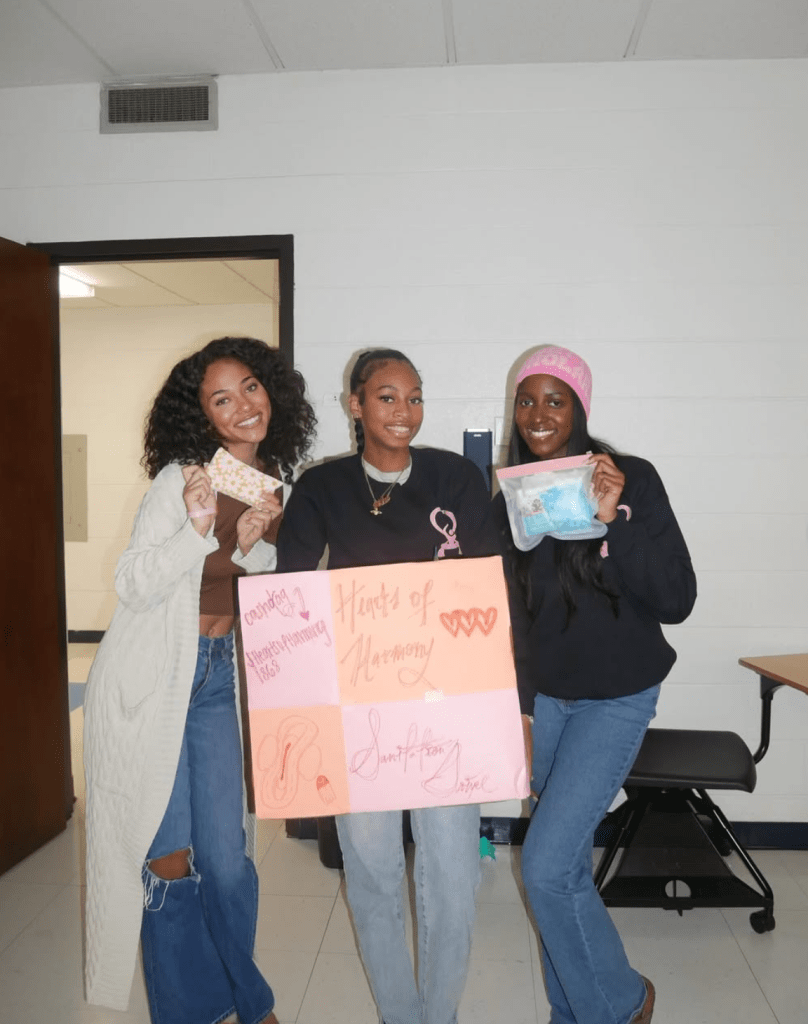 Three Hearts of Harmony co-founders smiling and holding a decorated donation board and menstrual health items during a campus service event.