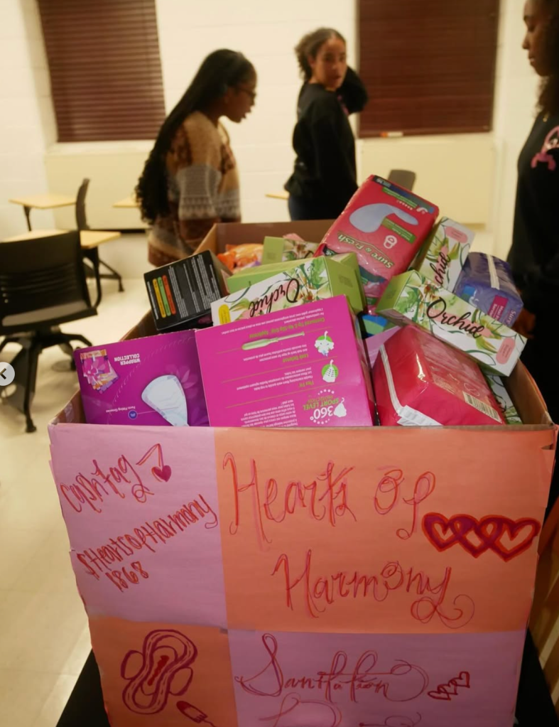 A large Hearts of Harmony donation box overflowing with pads and menstrual products as members sort supplies during a service initiative.