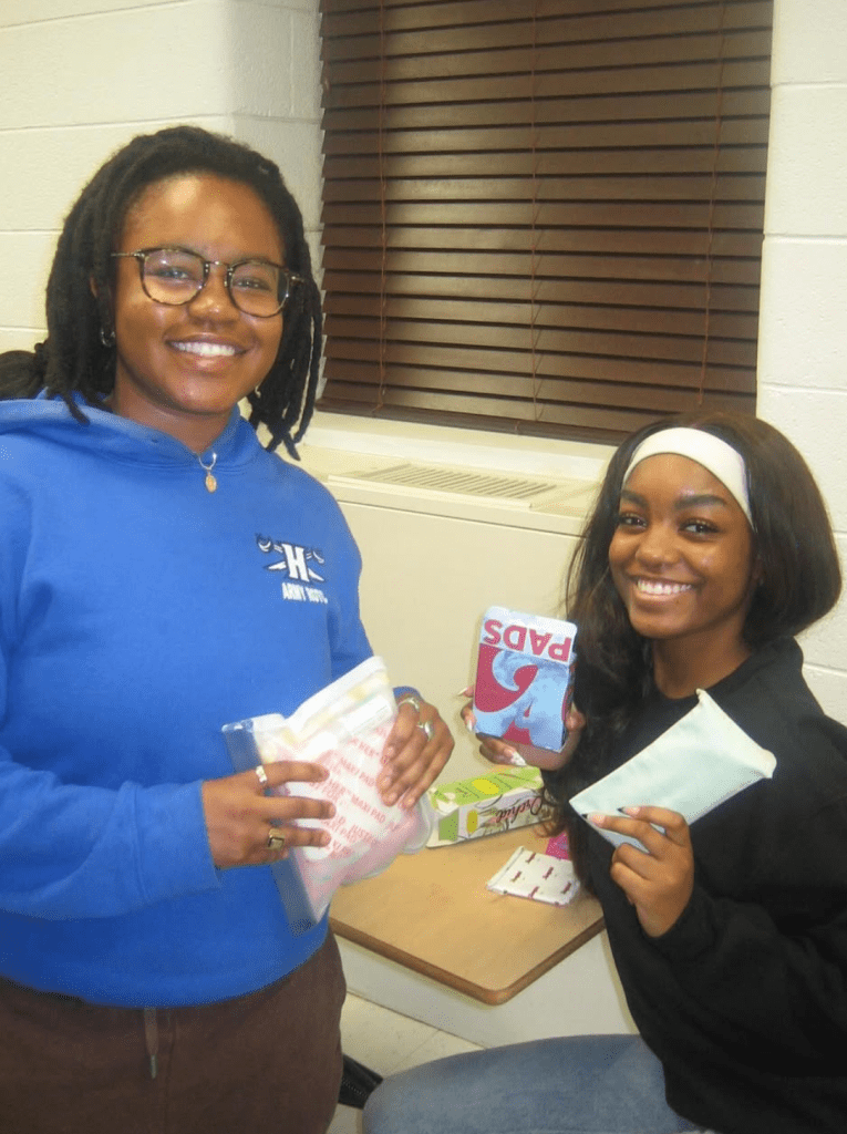 Two Hearts of Harmony volunteers smiling and holding menstrual products while assembling donation kits during a service night.