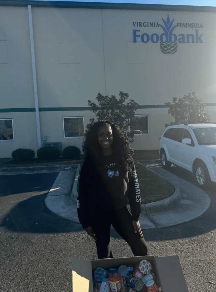 Miracle Gross standing outside the Virginia Peninsula Foodbank beside a large box of donated canned goods collected through the Fresh Start Fund initiative.