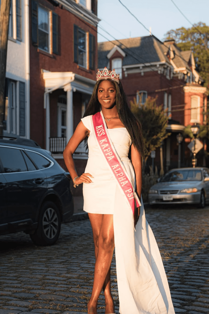 Miracle Gross poses for a professional outdoor portrait wearing her Miss Kappa Alpha Psi sash and crown, standing on a cobblestone street at sunset.