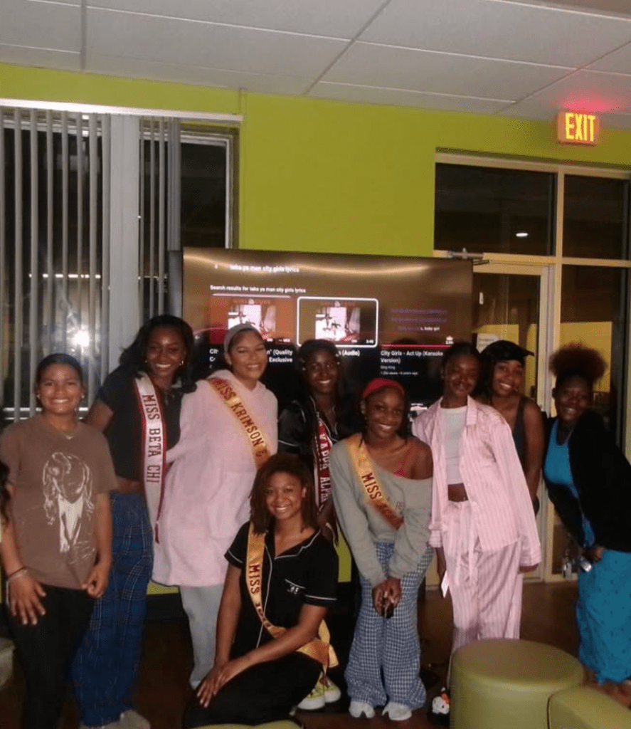 Miracle Gross and a group of campus queens pose together during a pajama-themed bonding night event, smiling in front of a screen displaying karaoke song options.