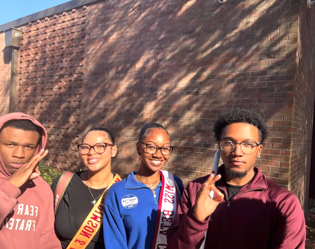 Kappa Kourt queens and fellow Kappa Alpha Psi Fraternity member smile outside a brick building while participating in community service at Mary S. Peake Elementary School. 