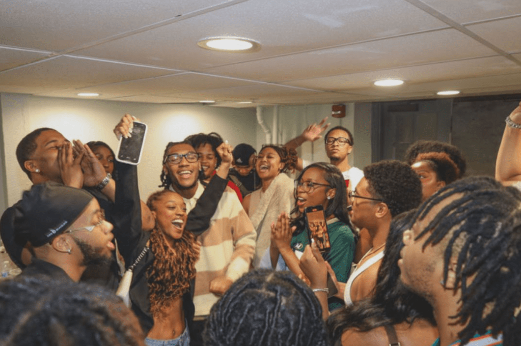 A group of students smiling, cheering, and raising their hands in celebration during a lively cast gathering for the BHX production Where Grace Finds Us.