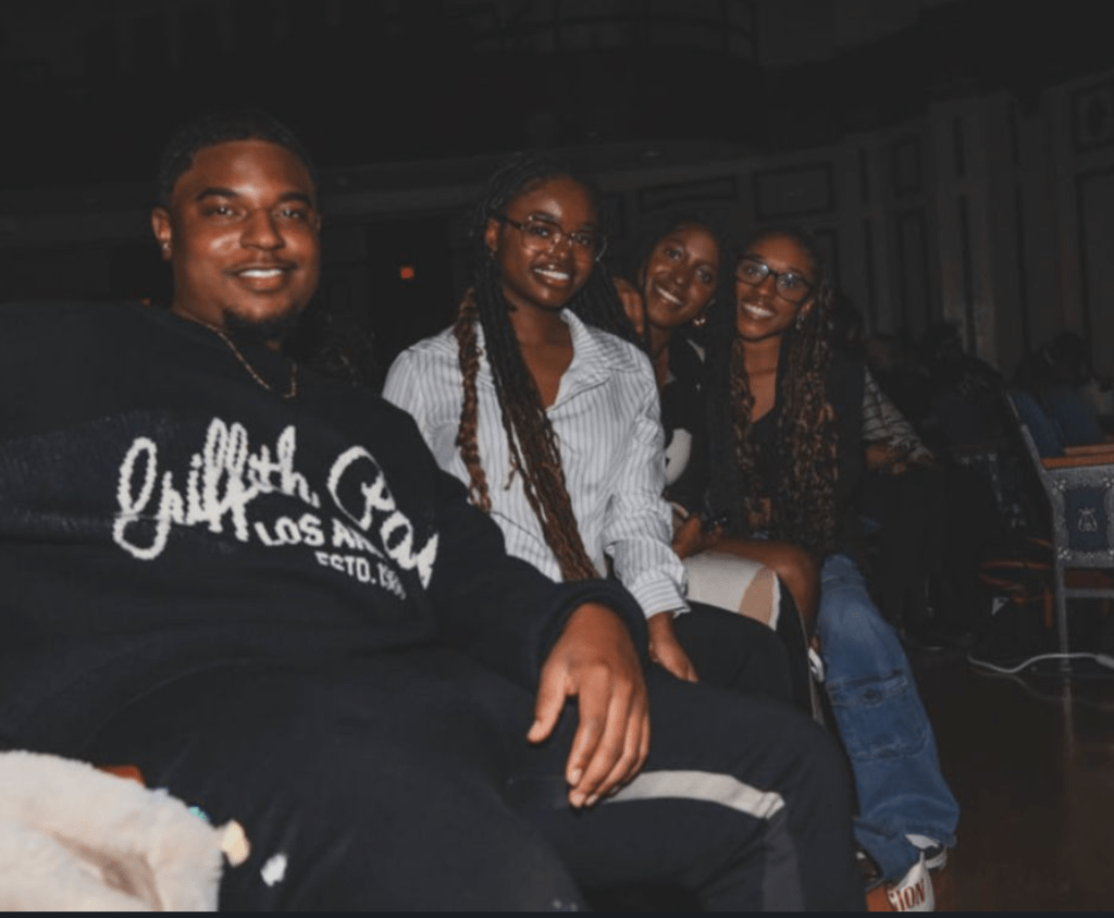 A group of four students sitting together in a dimly lit auditorium, smiling toward the camera during a campus event.